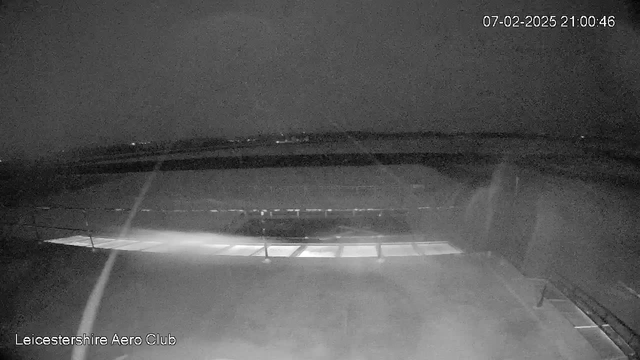 A dimly lit scene captured from a webcam, showing a view of a runway or open area at night. The image features a dark sky, with a few lights visible in the distance. A flat surface is present in the foreground, possibly a hangar or building, and there are faint lines indicating the edge of the structure. The overall atmosphere is very dark, creating a sense of night or low visibility conditions. A timestamp indicating the date and time is displayed in the upper right corner.