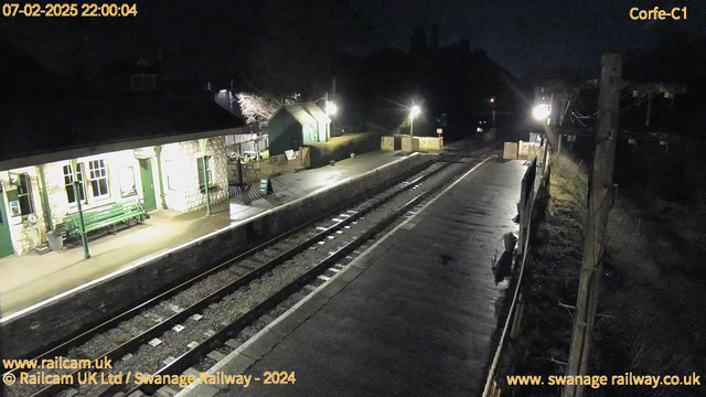 A dark station platform at night, with a stone building featuring large windows on the left side. There is a green bench along the platform, and a sign indicating "WAY OUT" partially visible. The platform is empty, with railway tracks running through the center, reflecting light from a nearby lamp. A wooden fence is visible in the background, and faint light emanates from a structure on the right side, creating a quiet, serene atmosphere.