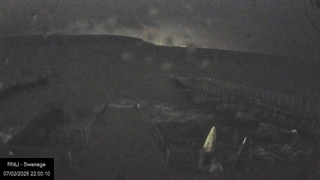 A dark scene captured from a webcam, showing a coastal area at night. The foreground features a partially illuminated boat with a white sail, positioned near a dock. In the background, faint lights from distant hills are visible, suggesting an elevated land area. The overall atmosphere is moody and somber, with a cloudy sky.