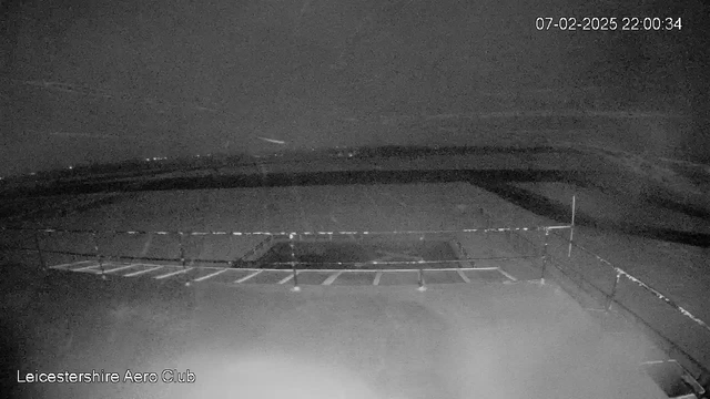 A low-light webcam image shows a dark scene with a small gravel or tarmac area in the foreground, bordered by a railing. The background appears to be open land or a runway, with faint outlines of trees or buildings in the distance, likely shrouded in darkness. The time stamp in the top right corner reads 07-02-2025, 22:00:34. The overall atmosphere is quiet and subdued, reminiscent of late evening or night conditions.