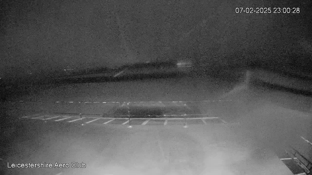 A dark webcam image showing a parking area at night. Several parking spaces are visible, with a blurred background indicating possible buildings or landscape features. The scene appears foggy or low visibility, with no clear details discernible. The date and time, 07-02-2025 23:00:28, are displayed in the top right corner. The bottom left corner shows the name "Leicestershire Aero Club."