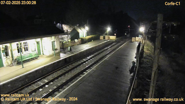 A dimly lit train station at night, featuring a long platform with two sets of railway tracks. On the left, there is a stone building with green details, including windows and a bench. There is a wooden fence in the background and a safety sign that says "WAY OUT." The right side of the image shows a utility pole with wires and a faint light in the distance. The scene is tranquil and empty, illuminated mostly by artificial lights.