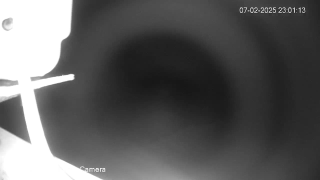 A black-and-white image from a webcam showing a dimly lit background with a blurred circular area. In the foreground, there is a partially visible object that resembles a light source or fixture on the left side. A cross-shaped white object extends horizontally across the bottom, with blurred edges indicating low clarity. The date and time are displayed in the top right corner.