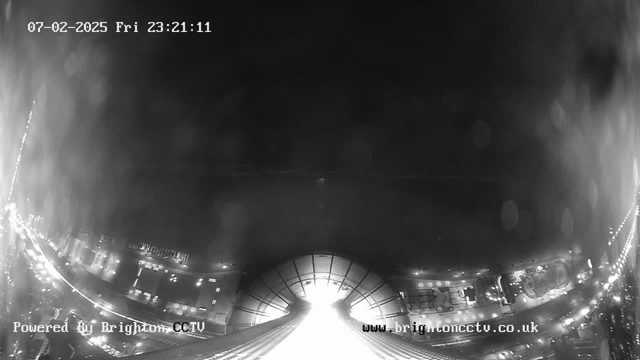 A black and white webcam image captures a view of a dark scene at night. The foreground features a large, circular structure that resembles a roof or dome. Below, there are faint outlines of buildings along the shoreline, with some lights visible reflecting off the water. The image has a timestamp indicating the date as February 7, 2025, at 23:21:11. The clarity is somewhat obscured by a mist or rain, creating a hazy effect across the scene.