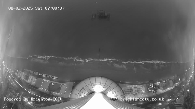 A black and white webcam image shows a view of the coastline with the ocean stretching toward the horizon. Waves are gently breaking along the shore, and a few structures, including a pier, are visible offshore. The sky appears overcast, creating a moody atmosphere. In the foreground, part of a curved roof is visible, along with buildings and walkways along the beach. The image contains a timestamp indicating it is early morning on February 8, 2025.