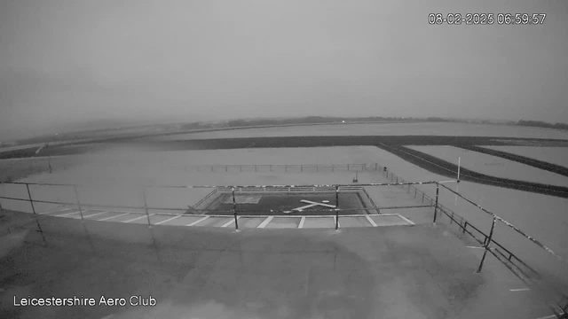 A black and white webcam image showing a wide, flat landscape with a low-lying area covered in water or fog. In the foreground, there is a helipad marked with a white “X.” The ground is mostly empty, and a safety railing is visible along the edge. The background features a flat horizon with no visible structures, suggesting a rural or open area, possibly an airstrip. The timestamp indicates the scene was captured early in the morning.