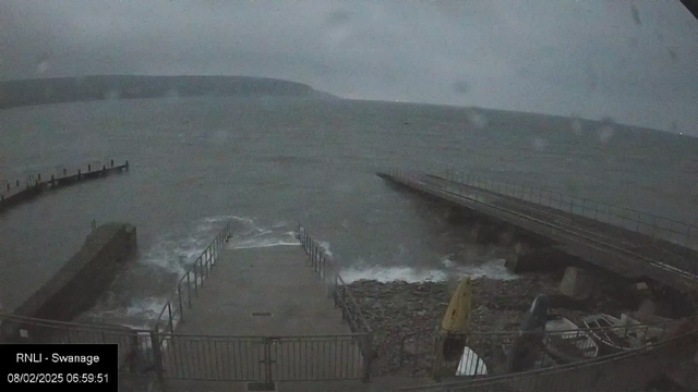 A dark, cloudy scene at a coastal area. The turbulent sea is visible in the foreground, with waves crashing against a rocky shore. A small concrete ramp leads down to the water, bordered by a metal railing. In the background, a wooden pier extends out into the water. The distant shoreline features a hilly outline under an overcast sky. The overall atmosphere appears gloomy, suggesting early morning or late evening.