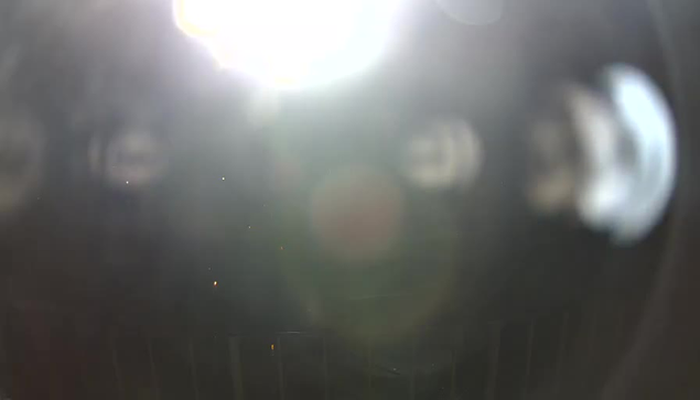 A bright light source is predominant in the image, creating a glare that obscures visibility. Surrounding the light, there are blurred circular shapes that may represent objects or reflections, while the background appears dark and unclear. A sense of motion or particles is suggested by faint spots scattered throughout the image.