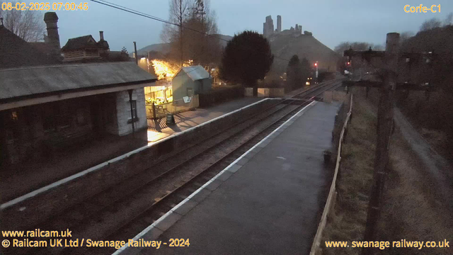 A dimly lit railway station scene at dawn features a stone building with a sloped roof on the left, partially obscured by trees. A few glowing lights suggest a small structure or café nearby. The tracks run parallel and are wet, likely from recent rain. In the background, a hill rises with remnants of a castle or tower structure on top, silhouetted against the gray sky. There are also utility poles along the right side of the image.