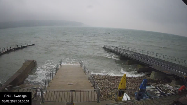 A cloudy seascape featuring a rocky shoreline, with stairs leading down to the water. Two piers extend into the water from the right side; one is straight and flat, while the other is slightly angled. There are several boats, including a yellow one, parked on the shore. The water is choppy with whitecaps, indicating rough seas. The scene is mostly gray and muted.