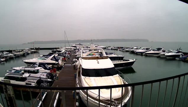 A view of a marina on a cloudy day, featuring various boats moored at the docks. Several white and blue boats are clustered on the water, with a few larger yachts in the foreground. The marina is surrounded by calm water, and there is a distant shoreline visible in the background. The atmosphere appears overcast, contributing to a grayish tone throughout the scene.