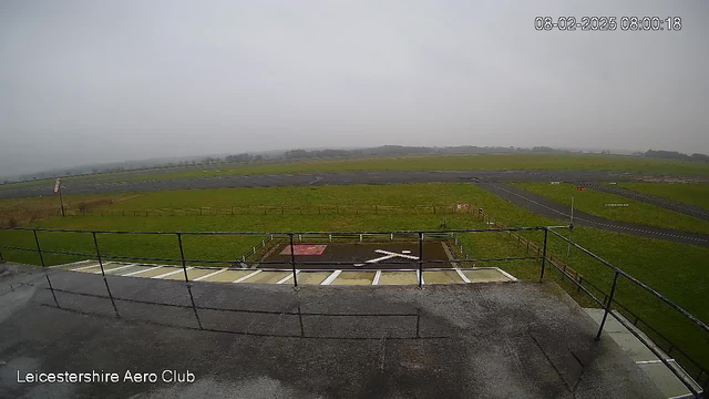 A view from a webcam showing a foggy landscape at the Leicestershire Aero Club. In the foreground, there is a railing and a section of grey pavement. Below the railing, there is a square area marked with a red surface and a white cross shape. The background features expansive green grass and a tarmac runway, stretching into the distance. The scene is overcast with a grey sky, and faint outlines of hills can be seen far away. The time displayed is 08:00:18 on February 8, 2025.