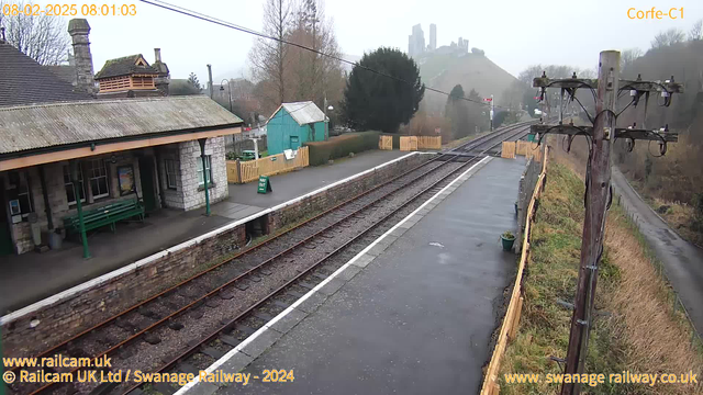 The image shows a railway station platform with a wet surface and several rail tracks visible. To the left, there is a stone building with a sloped roof and a covered waiting area containing a green bench. A green sign that reads "WAY OUT" is positioned in front of the building. In the background, there are trees and a misty hill with a castle-like structure on top. A wooden fence surrounds a grassy area, and there are several power lines and wooden poles along the right side. The scene appears cloudy and grey, suggesting overcast weather.