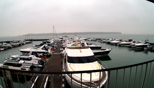 A marina scene with numerous boats docked in the water. The foreground features a white motorboat with a covered cockpit, while several other boats in various colors are arranged along the docks. The water is calm and reflects the cloudy, gray sky. In the background, a distant shoreline is visible, partially obscured by haze. The overall atmosphere is quiet and serene.