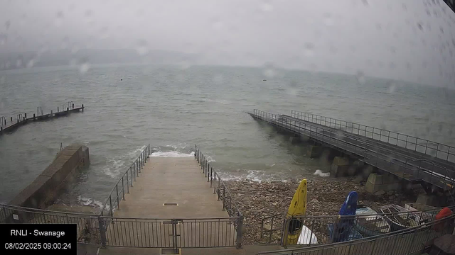 A foggy scene by a coastline shows a small cement ramp leading down to the water, surrounded by railings. On the left, there are two piers extending into the sea, with several docking posts visible. In the foreground, some boats are stored on the rocky shore, including a yellow kayak and other colorful covers. The sky appears gray and overcast, indicating rainy weather. Small waves are visible at the water's edge.