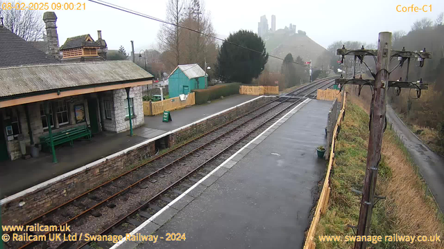 A view of a railway station platform on a cloudy day. On the left, there is a stone building with a sloped roof and several windows. A green bench is positioned on the platform. In the background, a light blue wooden building and a yellow wooden fence are visible. The platform has exposed railway tracks running through it, leading into the distance. On the right, there are utility poles with wires and a glimpse of a hilly landscape with ruins in the background. The ground is damp, indicating recent rain.