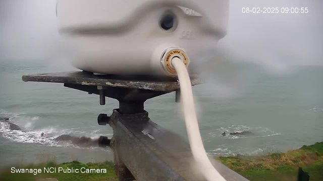 A close-up view of a white camera mounted on a metal support, with a flexible cable connecting it to a nearby surface. The background shows an overcast sky and the sea, with waves crashing against rocky formations along the coast. The image is slightly blurred due to moisture on the lens, giving it a hazy appearance.