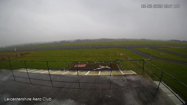 A view from a webcam at Leicestershire Aero Club, showing a cloudy, overcast sky. The foreground features a railing, and a paved area with a large white 'X' marking on grass below. In the background, there is a wide green field and a grey tarmac runway, with a few distant trees and structures partially obscured by the fog. The date and time display is in the top right corner, indicating it is early morning.