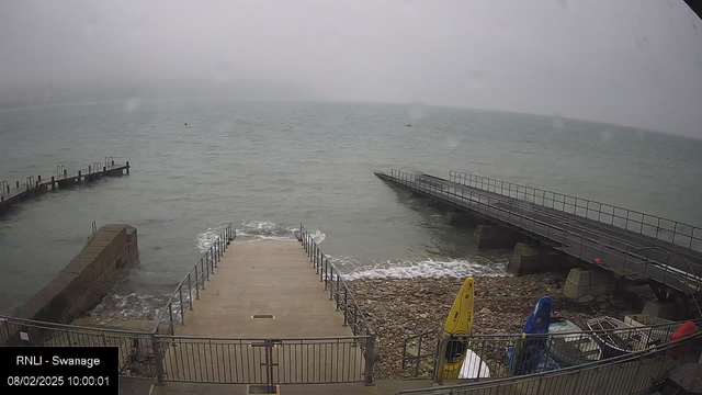 A cloudy scene over a body of water with gentle waves. In the foreground, there is a wide, concrete ramp leading down to the water’s edge, bordered by a metal railing. On the left side, a small jetty with several posts extends into the water. To the right, another jetty is visible, also leading into the water. In the lower right corner, there are various colored canoes and equipment stored near the edge, with some rocky ground beneath them. The atmosphere is grey and misty, indicating overcast weather.