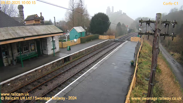 A view of Corfe Castle railway station, featuring a weathered stone structure with a tin roof on the left. A green bench is visible under the roofed area. The platform is lined with gravel tracks, leading into the distance. There is a green sign that reads "WAY OUT," and a wooden fence in the background. Additionally, the ruins of Corfe Castle can be seen on a hill in the background, partially obscured by fog. A power pole with wires stands on the right side of the image. The scene is dreary with overcast skies.