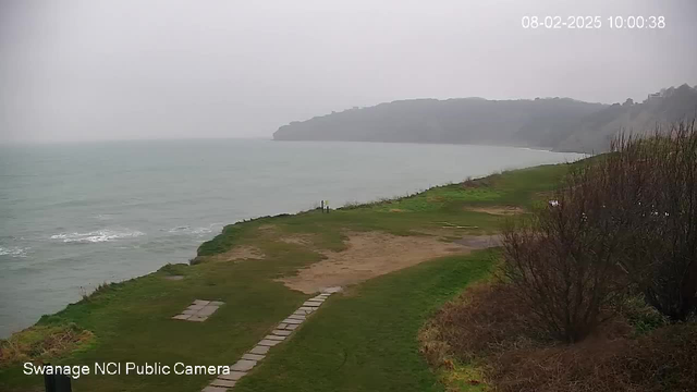 A cloudy, overcast scene showing a coastline. The calm sea is visible with gentle waves. In the foreground, there is a grassy area with a dirt path made of stone slabs leading toward the water. To the right, there are patches of sparse vegetation and shrubs. The setting appears tranquil and somewhat misty, indicating a dull day.