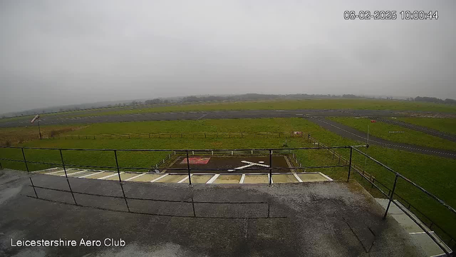 A view from a webcam showing a flat grassy area, likely an airfield, under a cloudy sky. In the foreground, there's a railing and a flat surface with a large white 'X' marking. The grassy area extends into the distance, with a black runway and some dirt paths visible. A windsock is positioned on the left, indicating wind direction. The weather appears overcast, suggesting a dreary atmosphere. The timestamp at the top shows the image was captured at 10:00 on February 8, 2025.