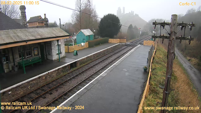 A foggy scene at a train station with a stone building on the left. The building has a sloped roof and a green bench outside. There are some trees and a small green shed in the background. The platform has empty train tracks leading away, and there is a sign that reads "WAY OUT." A wooden fence runs along the platform, and in the distance, a castle ruins can be seen shrouded in fog. The ground is wet, indicating recent rain.