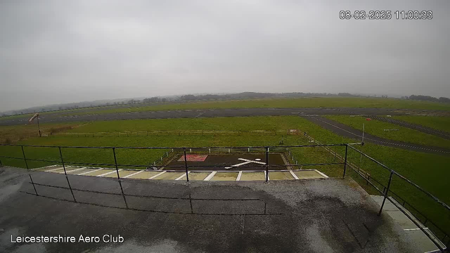 A view from a webcam at the Leicestershire Aero Club, showing a large grassy area with a partially visible runway and taxiway. There is an "X" marking on the ground, indicating a no-landing zone, and some fencing surrounds the area. The sky is overcast and gray, giving a dull appearance to the landscape. The image captures the edges of a balcony or roof in the foreground, with moisture visible on the surface.