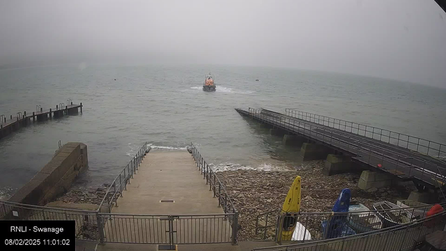 A foggy coastal scene featuring a small, orange rescue boat navigating through calm waters, moving away from a concrete pier. On the left, a set of stairs leads down to the water, bordered by a railing. In the foreground, there are several boats and kayaks of various colors, including a yellow kayak and a blue cover. The atmosphere is muted due to the fog, with visibility limited. The date and time are displayed in the lower left corner, showing "08/02/2025 11:01:02."