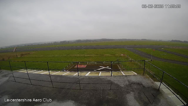 A view from a balcony overlooking a grassy airfield. The sky is overcast with a grayish hue. In the foreground, there is a flat surface with a railing. Below, a grassy area includes a marked landing zone with a prominent white "X" shape. Various patches of grass and a few runway markings can be seen in the background, along with a distant line of trees. The scene appears calm and empty, typical of an aviation facility. The timestamp in the corner indicates the date and time of the image capture.