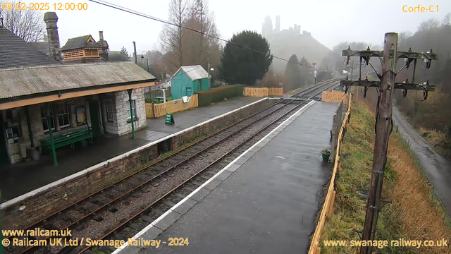 A view of Corfe Castle railway station on a cloudy day. The station features a stone building with a sloped roof and decorative chimney. There is a green wooden bench on the platform, and a wooden fence borders the area. A green shed and other structures are visible in the background, along with a signal pole on the right side. The railway tracks run parallel to the platform, which has a damp surface, and in the distance, mysterious hills and the outline of Corfe Castle can be seen shrouded in mist.