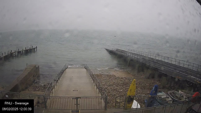 A foggy scene at a waterfront with a rocky shoreline. In the foreground, there is a staircase leading down to the water, bordered by a metal railing. To the left, a wooden pier extends into the water, with several posts visible. The water is calm but appears gray under overcast skies. On the right side, there are two kayaks, one yellow and one blue, beside a storage area with more boats partially covered. The scene is damp, likely due to rain, as droplets are visible on the camera lens.