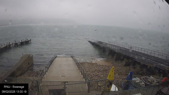 A foggy seaside scene shows a partially submerged ramp leading down to the water, flanked by a rocky shoreline. To the right, there are several boats partially covered with blue and red tarps. A long, weathered pier extends into the calm gray water, and another pier can be seen at the left, with several posts jutting out. The overcast sky appears dull and misty, suggesting rain. Overall, the atmosphere is tranquil but hazy, with limited visibility of distant objects.