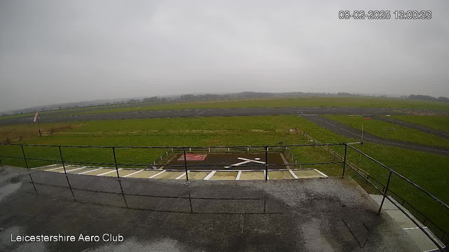 A view from a raised platform overlooking an airfield on a cloudy day. The ground is primarily green grass, with a dark asphalt runway visible in the distance. A short fence lines the perimeter of the airfield, and a red sign is partially visible near the runway. The sky is overcast with gray clouds. The foreground includes a railing along the platform, and the image appears to be wet, suggesting recent rain.