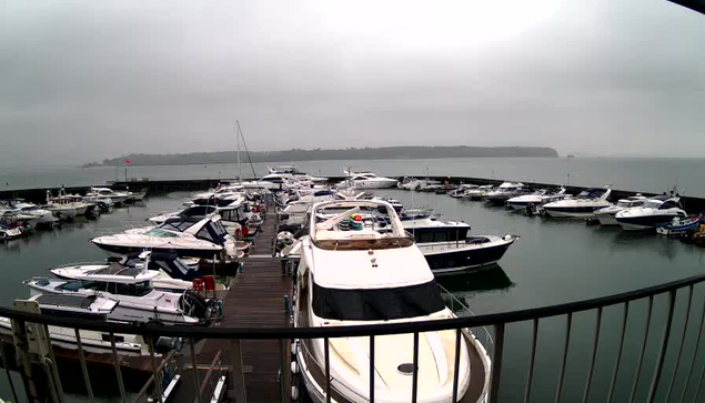 A marina filled with various boats docked along a wooden pier. The water is calm and reflects the cloudy, overcast sky. Some boats are larger yachts, while others are smaller motorboats. In the background, there is a distant shoreline partially obscured by mist, and a flag is visible waving on the pier. The scene conveys a tranquil, subdued atmosphere.