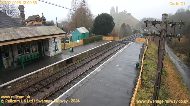 A train station scene on a cloudy day, featuring a stone building with a sloped roof and overhanging eaves. There is a green bench on the station platform, and beyond, two sets of railway tracks lead off into the distance. On the left, a wooden pole with electrical wires supports multiple devices. In the background, a foggy hill rises, displaying remnants of castle ruins at the top. A small turquoise shed is visible along with a wooden fence demarcating an area with signage saying "WAY OUT." The ground appears wet, indicating recent rain.