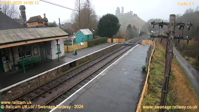 A railway station scene on a cloudy day. The foreground features a platform with a green bench and a stone wall beside the railway tracks. Perpendicular to the tracks, a wooden post with electrical lines stands on the right. In the background, a misty hill is visible with ruins or a castle-like structure. A small turquoise building is partially obscured by trees, and there is a wooden fence indicating an exit path. The ground is wet, indicating recent rain.