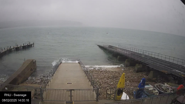 A view of a harbor under overcast skies, with choppy water. A series of wooden piers and ramps lead down towards the water, with some visible waves lapping at the shore. In the foreground, there is a rocky shoreline with a couple of yellow and blue kayaks positioned by the railing. The overall scene conveys a grey, misty atmosphere, typical of a cloudy day at the seaside.