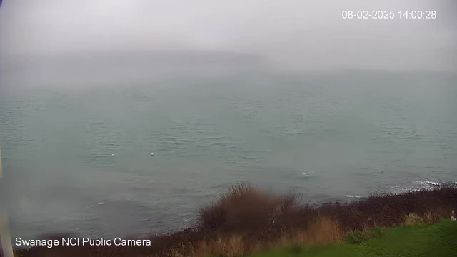 A foggy coastal scene featuring the sea with gentle waves. The horizon is obscured by mist, and the water appears calm with a light greenish hue. In the foreground, there is a patch of land with some brown shrubbery and grasses. The left side of the image is mostly hidden by mist, contributing to the overall hazy atmosphere. The date and time are displayed in the top right corner.