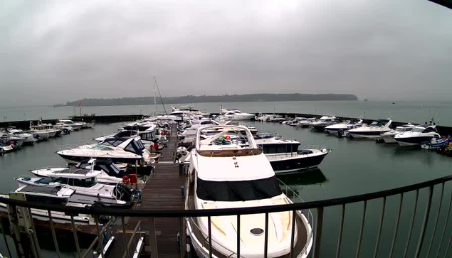 A view of a marina on a cloudy day, featuring a dock filled with various white and blue boats. The water is calm and reflective, and in the background, land is visible on the horizon. The scene conveys a tranquil but overcast atmosphere.