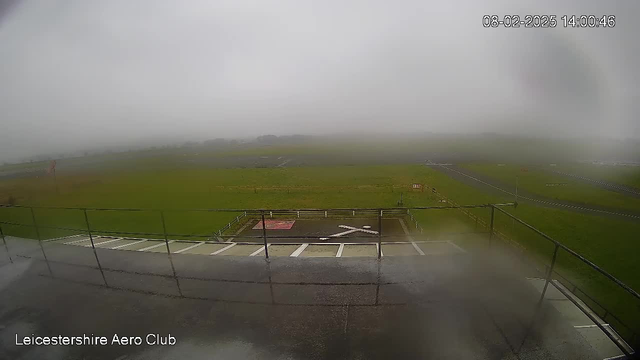 A foggy scene from a webcam at Leicestershire Aero Club. The foreground shows a wet balcony railing with gray concrete. Below, there is a grassy area with a large white "X" marking. In the distance, the grass continues into a blurred, foggy background, making it difficult to see further details. The sky is overcast and gray. The timestamp in the corner shows the date and time.