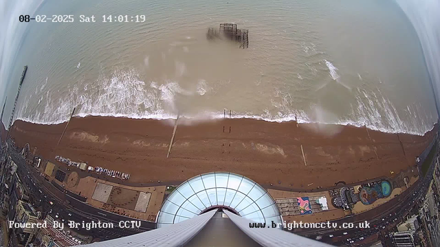 A view from a high vantage point overlooking the coastline. The image shows sandy beach with small waves lapping against the shore, and an old pier partially submerged in the water. The beach is bustling with people, and various colorful structures and attractions are visible near the shoreline. In the foreground, the glass dome of a building is prominently featured. The sky is overcast, suggesting a cloudy day. The timestamp indicates it is February 8, 2025, at 14:01.