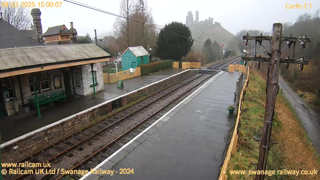 A train station platform is shown, featuring a stone building with a sloped roof, green benches, and metal poles. It is a cloudy day with a misty atmosphere. In the background, a hill rises with a castle or ruins at the top. The track runs parallel to the platform, which is wet from rain, and there are yellow and green signs visible. A wooden fence borders the area.