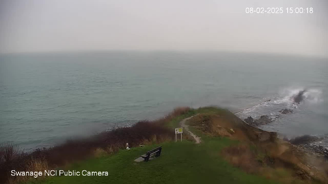 A coastal scene shows a grassy area with a wooden bench facing the ocean. The water is calm and reflects a grey sky, indicating overcast weather. There is a path leading from the bench towards a sign, and rocky formations are visible where the water meets the shore. The atmosphere appears misty, with limited visibility in the distance.