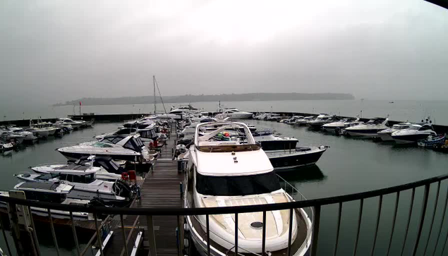 A cloudy and overcast day at a marina, featuring many boats docked in the water. The scene shows a variety of yachts and smaller vessels moored along wooden docks, with a railing visible in the foreground. The water appears calm with reflections of the boats and sky. A distant shoreline is partially visible in the background, shrouded in mist.