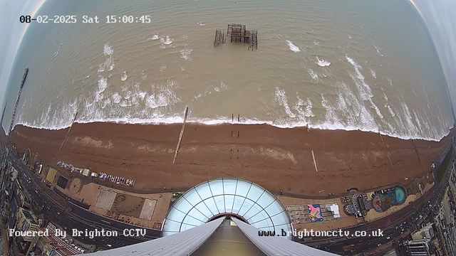 Aerial view of a sandy beach with gentle waves lapping at the shore. In the foreground, a circular glass structure is visible, likely part of an observation deck. The beach is lined with a few buildings, some amusement rides, and scattered seating areas. In the distance, an old pier structure juts out into the water. The sky appears cloudy with hints of blue. The image has a timestamp indicating the date and time, and a watermark in the corner.