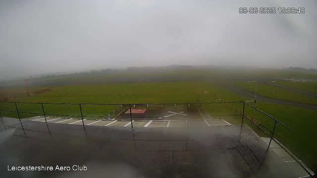 A blurry, foggy scene depicting an open field with a few visible runway markings and a small red landing area. A railing is visible in the foreground, and the landscape appears green, likely grass, under overcast skies. Visibility is low due to fog, obscuring distant objects.