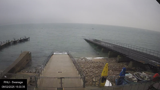 A foggy seaside view featuring a calm sea with visible ripples. In the foreground, there is a concrete ramp leading down to the water, surrounded by a metal fence. To the left, a wooden pier extends into the water with several posts. On the right, there are boats and kayaks in various colors, including yellow and blue, resting on the shore. The atmosphere is overcast, creating a muted color palette.