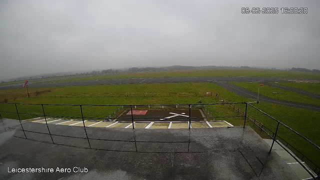 A view from a webcam showing a cloudy sky over an airfield. In the foreground, there is a railing with a concrete surface, and a logo on the ground resembling an X shape. The background features a green grassy area and an asphalt runway. The airfield extends into the distance, where low hills are barely visible through the mist. A windsock is positioned to one side, indicating light wind conditions. The date and time at the top right corner show February 8, 2025, at 4:00 PM.