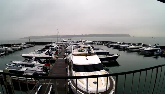 A marina filled with various boats and yachts is seen from a webcam perspective. The boats are docked in an orderly manner, with some white boats prominent in the foreground and a row of darker-colored boats in the background. The water is calm and reflects the overcast sky, which is gray and foggy. A wooden dock is visible in the lower part of the image, and the horizon features a distant landmass shrouded in mist.